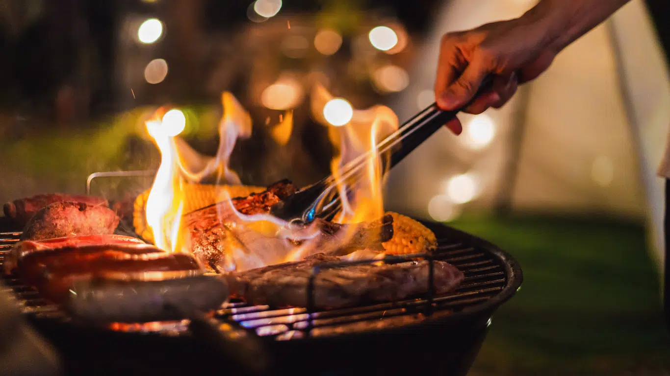 Et si le café devenait la star de vos barbecues d’été ? Photo, de nuit, avec des lumières scintillantes en fond, d'une main s'occupant d'un barbecue